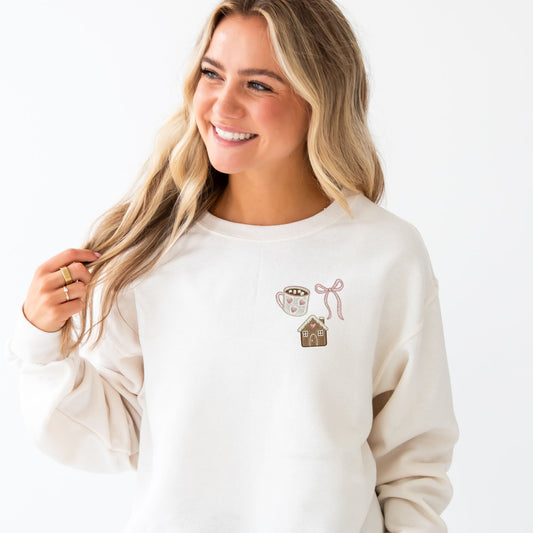 Woman wearing a white sweatshirt with a mug, bow, and gingerbread house embroidered house.
