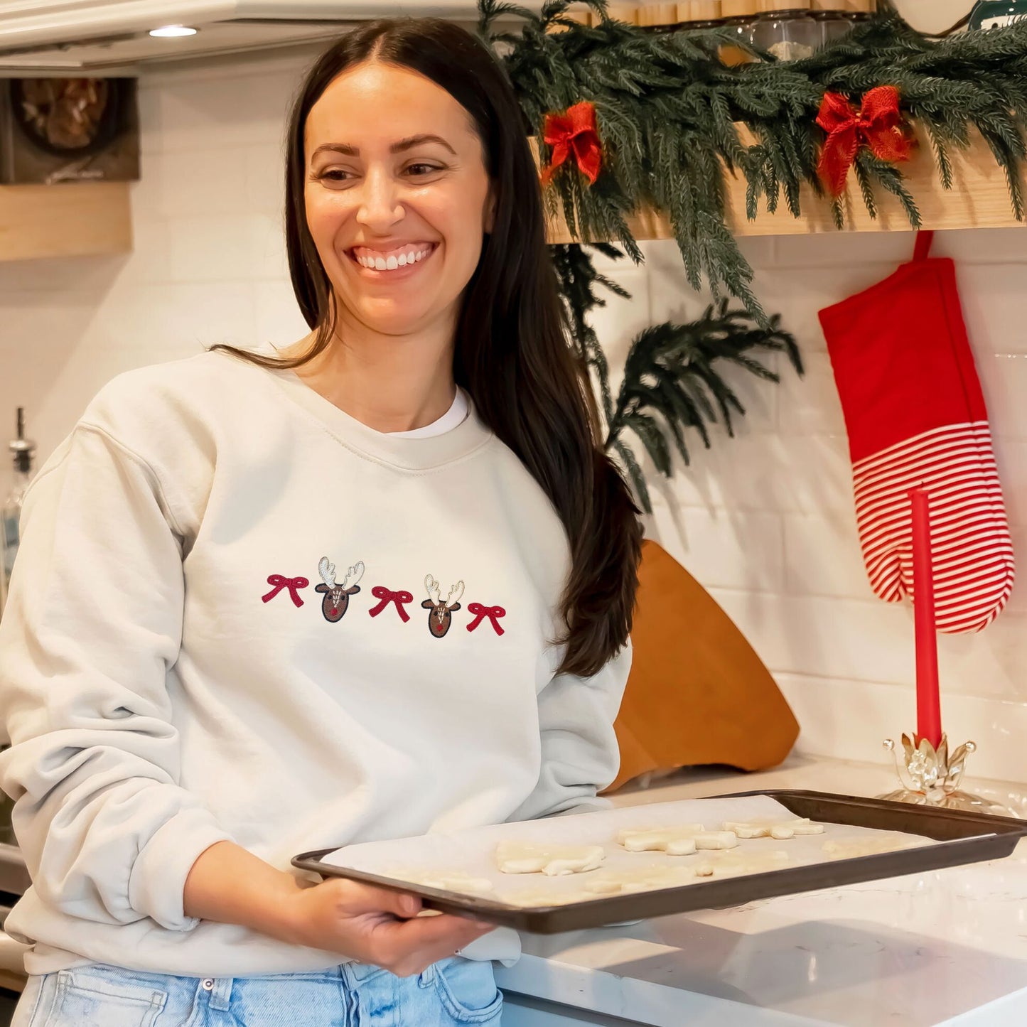 woman baking in the kitchen wearing a sand crewneck sweatshirt with custom bow/reindeer embroidered design across the chest