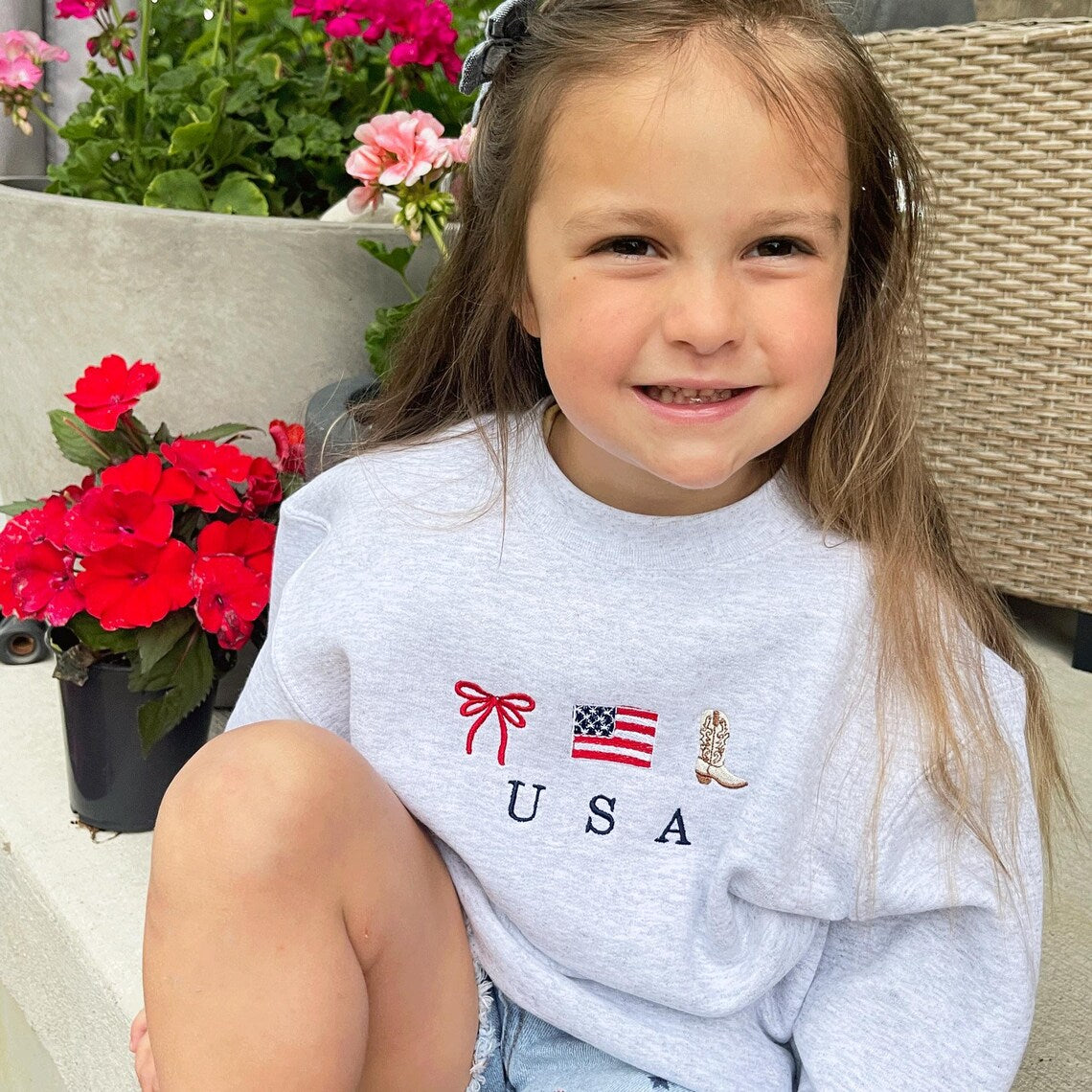 little girl sitting on front porch next to colorful flowers wearing jean shorts and a cute ash crewneck sweatshirt with an embroidered usa design featuring a double loop bow, american flag, and cowboy boot