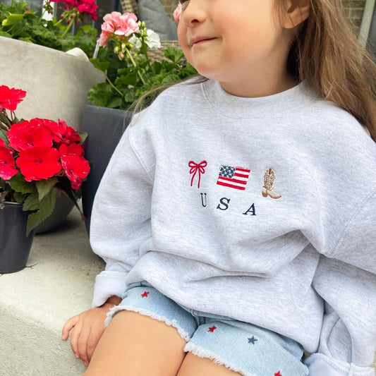 close up of little girl sitting on front porch next to colorful flowers wearing jean shorts and a cute ash crewneck sweatshirt with an embroidered usa design featuring a double loop bow, american flag, and cowboy boot