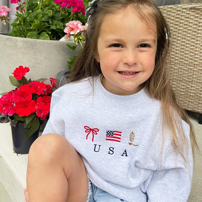 little girl sitting on front porch next to colorful flowers wearing jean shorts and a cute ash crewneck sweatshirt with an embroidered usa design featuring a double loop bow, american flag, and cowboy boot