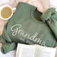 Flat lay of a military green crewneck sweatshirt styled with coffee and a book. On the center chest of the crewneck is embroidered Grandma est 2020 in white thread.