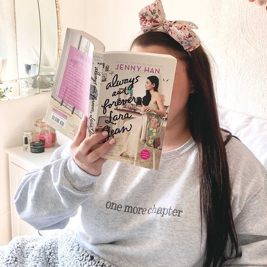 girl lounging in bed with a book wearing "one more chapter" booklover crewneck