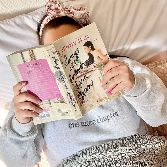 young woman wearing "one more chapter" sweatshirt while reading a book in bed