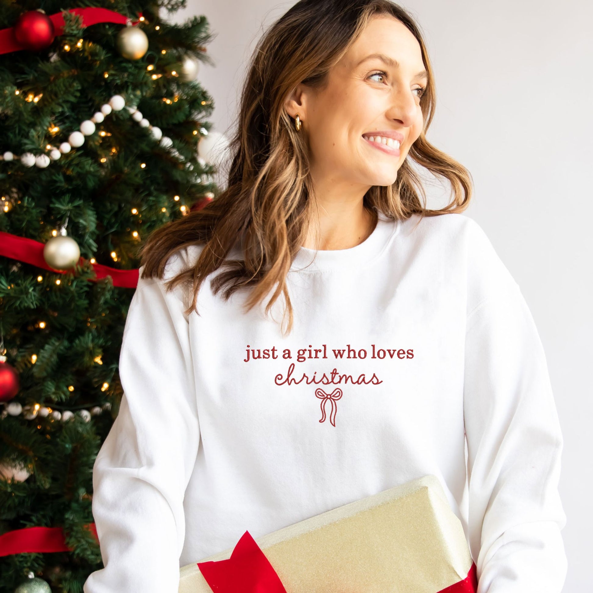 Woman wearing a white sweatshirt with 'just a girl who loves Christmas' text, standing next to a decorated Christmas tree.