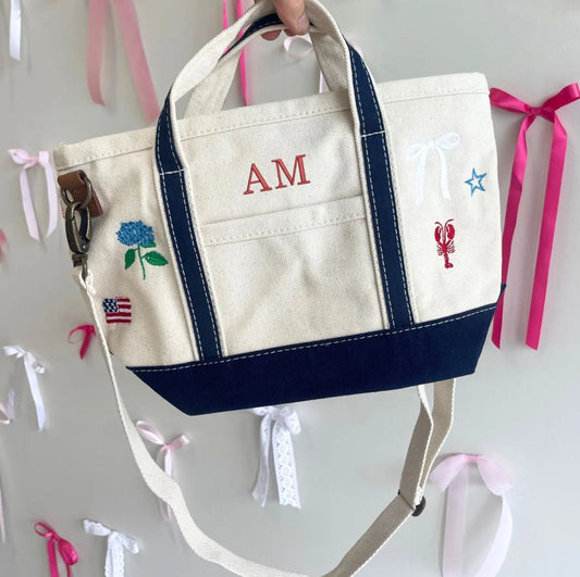 Canvas tote bag with embroidered designs and initials, held against a light background with ribbons.