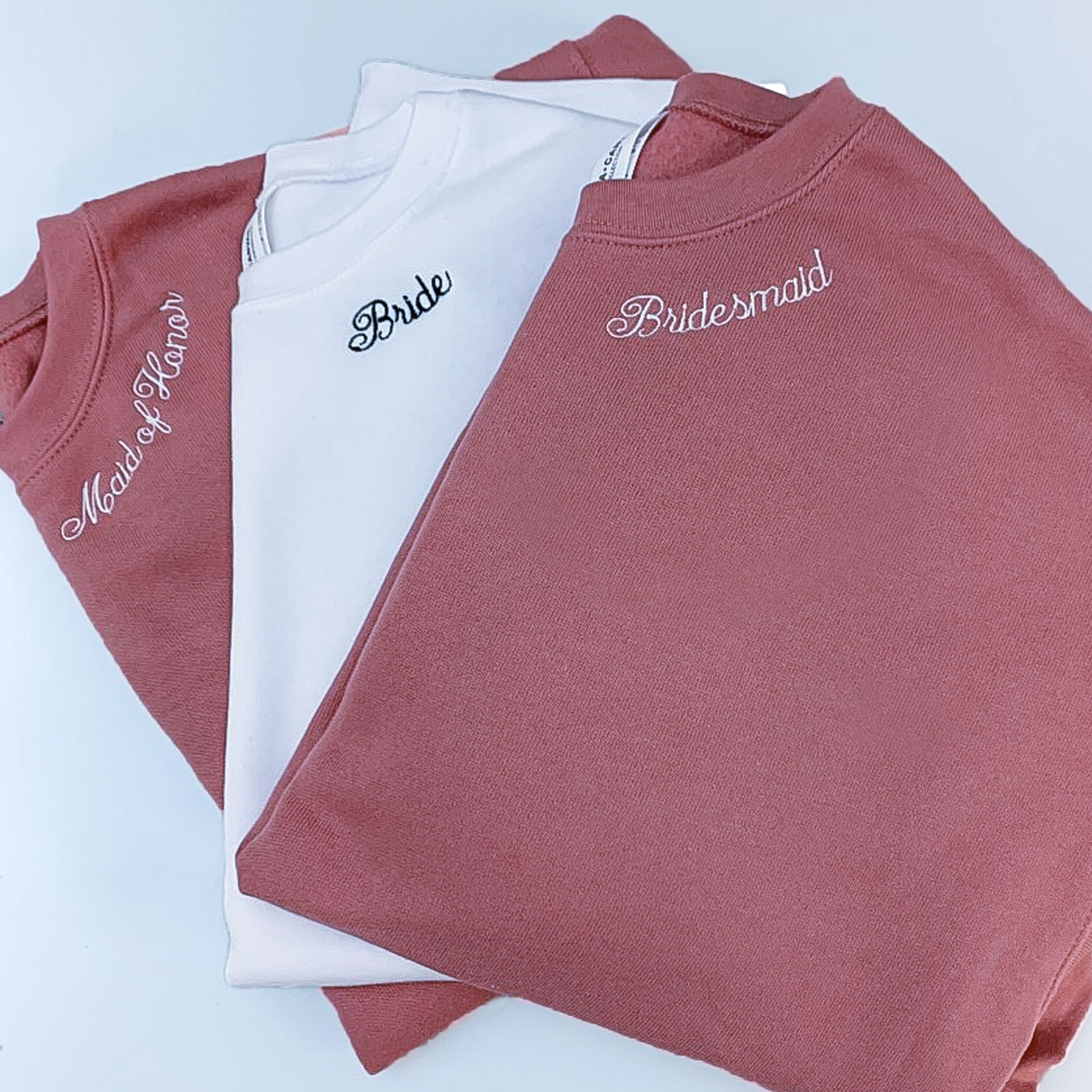 flat lay of two mauve crewneck sweatshirts and and a white crewneck sweatshirt with neckline bride and bridesmaid embroidery in white and black threads 