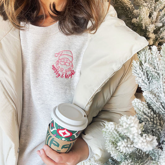 woman wearing an ash gray sweatshirt with a cute pink and red embroidered santa with merry and bright underneath