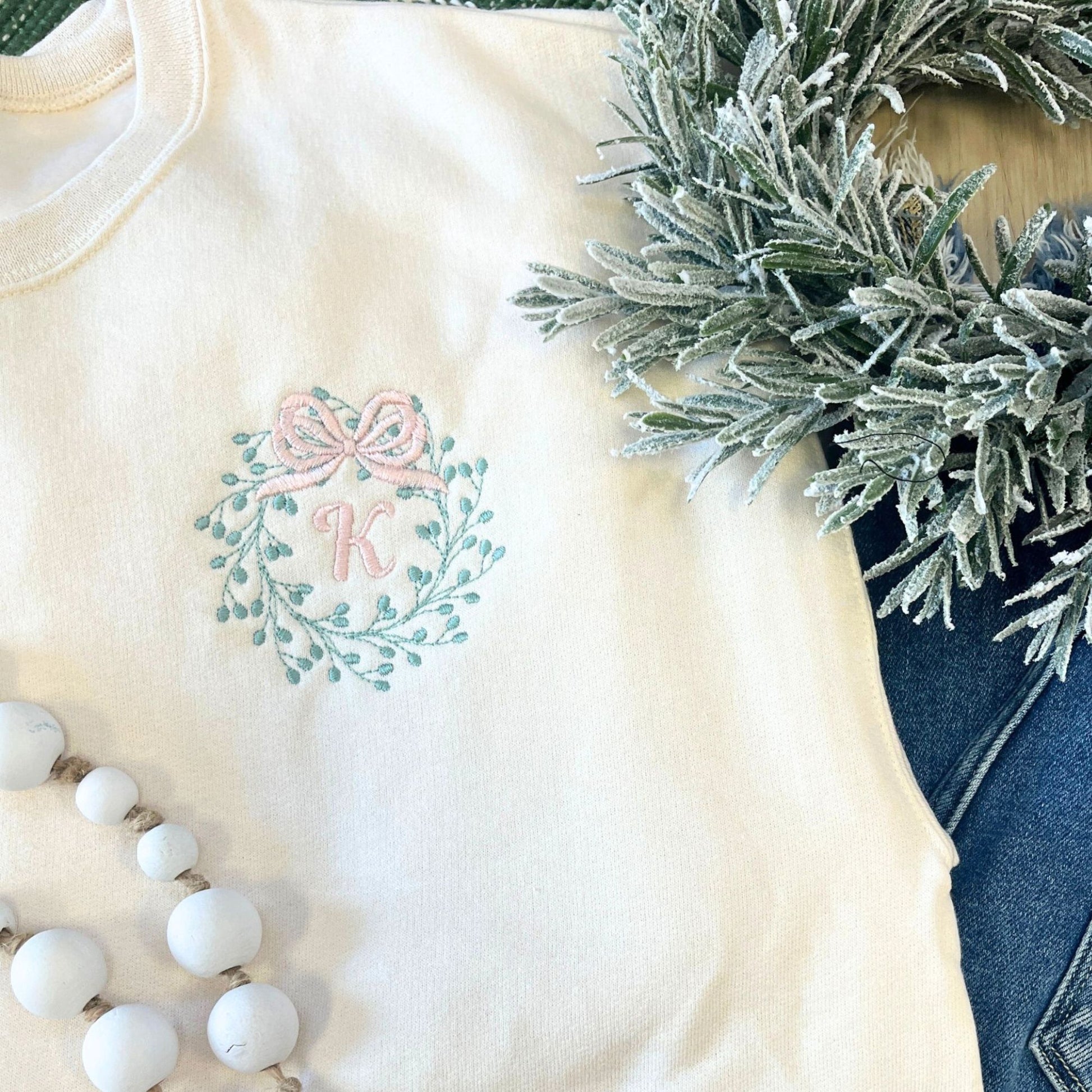 sweet cream shirt with embroidered design featuring a pink bow and letter 'K', next to a wreath and beads.