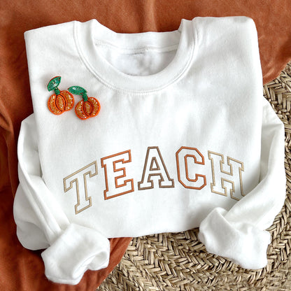 styled flat lay photo of a white sweatshirt with embroidered all caps TEACH in alternating fall colors.