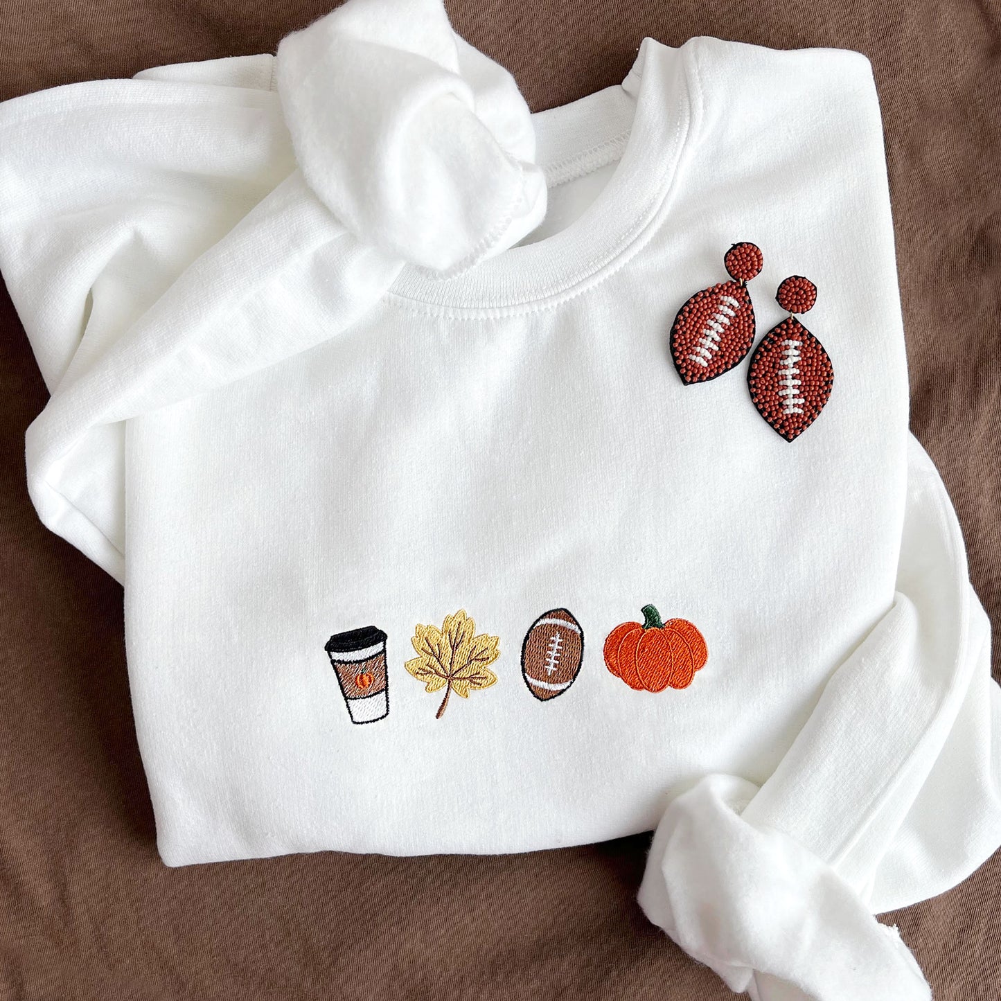 styled flat lay photo of a white crewneck sweatshirt with embroidered coffee, leaf,  football, and pumpkin design across the chest. 