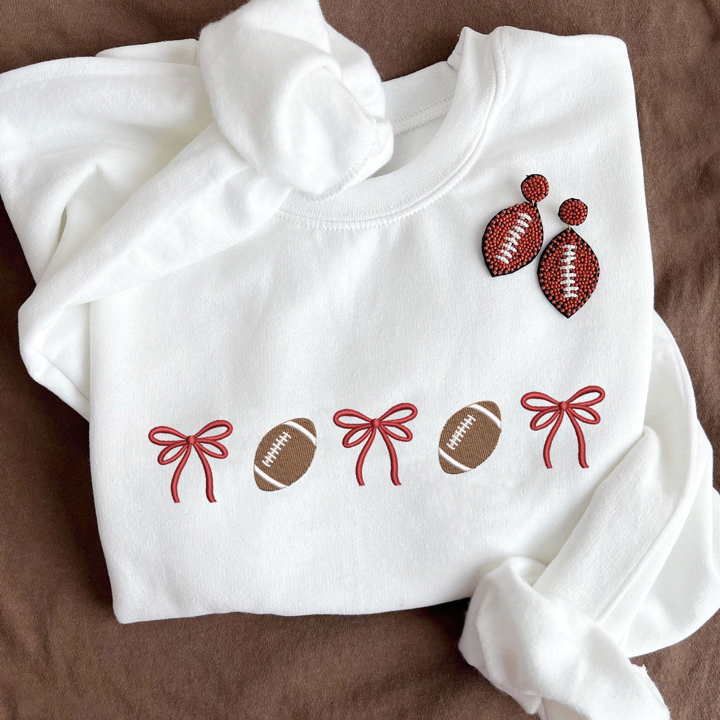 flat lay of a white crewneck sweatshirt with embroidered alternating bows and footballs design across the chest. The bow are embroidered in red thread.