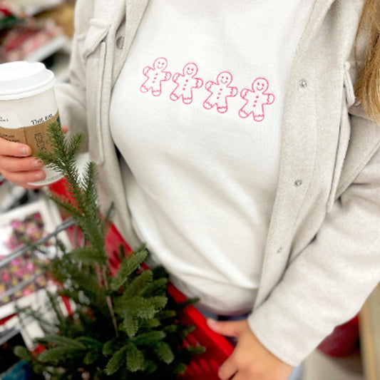 woman wearing a white crewneck sweatshirt with 4 pink embroidered gingerbread men across the chest. the woman is shopping for Christmas decor and holding a Starbucks cup.