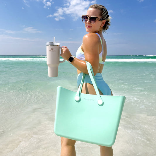 girl on a beach with the ocean in the background, holding a mint green large tote in the crook of her arm and holding a large water bottle