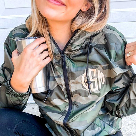 woman wearing a full zip windbreaker jacket with a forest green camo pattern and embroidered personalized monogram in a natural thread.