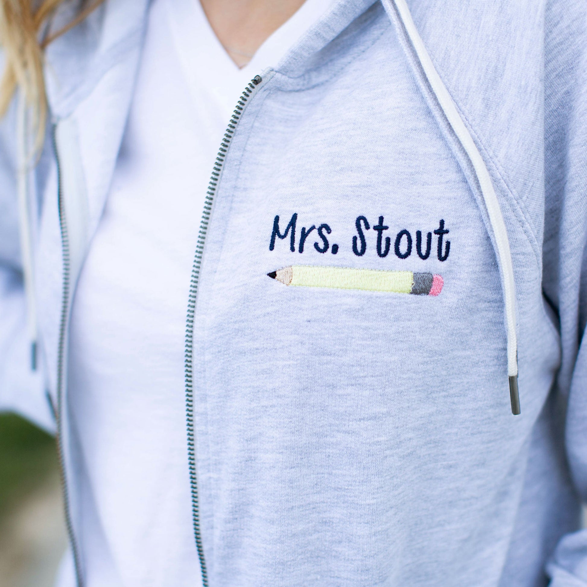 close up of educator's name and pencil embroidered on a grey zippered jacket with white and grey double string