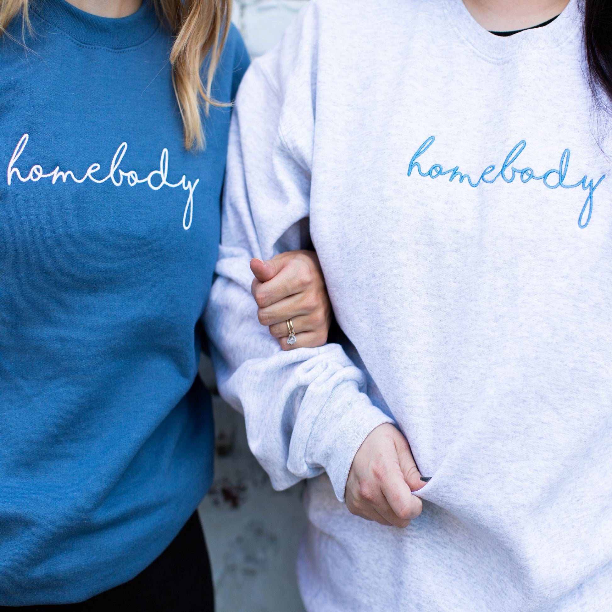 two girls standing side-by-side wearing crewneck pullovers one is blue with homebody embroidered in white thread across the chest and the other is ash gray with homebody embroidered in blue thread across the chest 