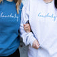 two girls standing side-by-side wearing crewneck pullovers one is blue with homebody embroidered in white thread across the chest and the other is ash gray with homebody embroidered in blue thread across the chest 