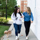 a blonde woman and brunette woman walking a dog side by side wearing matching homebody embroidered sweatshirts in light gray and blue