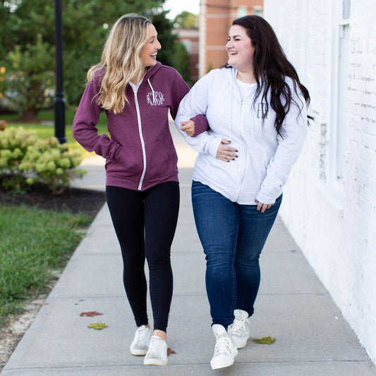 Two girls walking side by side. Blonde wearing a maroon full zip jacket with white monogram embroidery and brunette wearing ash gray full zip jacket with blue monogram embroidery 