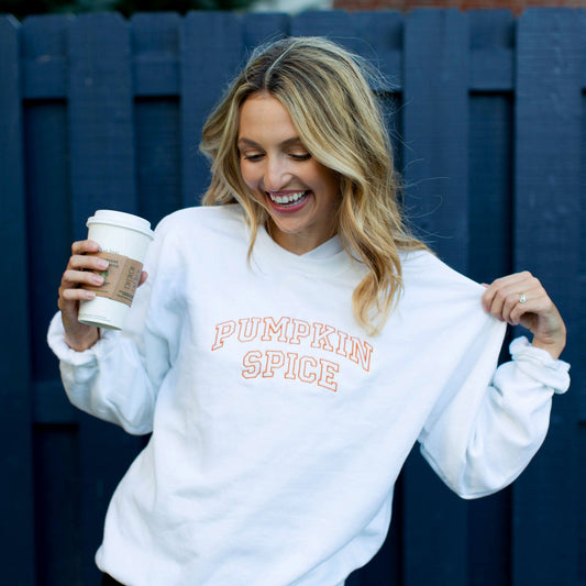 girl with travel coffee cup wearing a white sweatshirt that has pumpkin spice embroidered in orange thread across the front