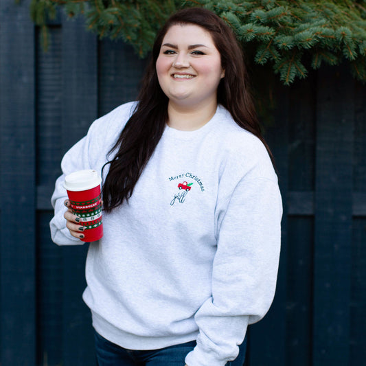 girl holding a travel coffee cup wearing a ash pullover crewneck sweatshirt with merry christmas yall and a small red truck embroidered on the left side