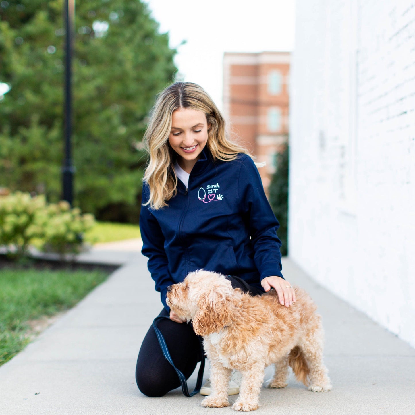 Woman kneeling next to dog wearing a navy polyester full zip jacket with embroidered pink stethoscope, paw print, and technician's name