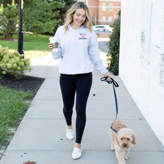 Woman wearing an ash gray pullover sweatshirt with hello pumpkin and a mini pumpkin embroidered on the left chest walking her dog and carrying a cup of coffee