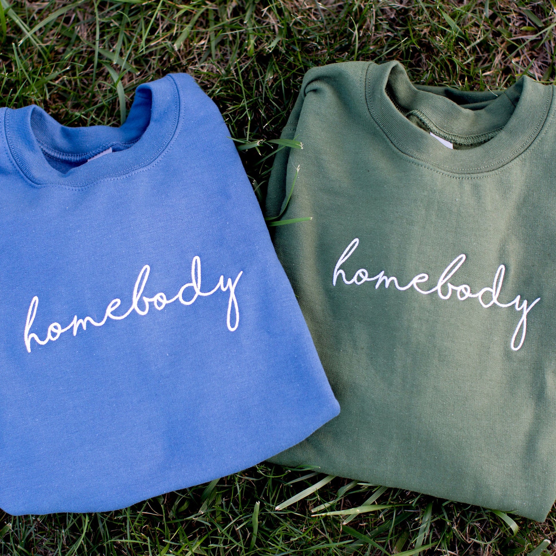 indigo and olive green sweatshirt with homebody embroidered in white thread and a script font folded and laying on grass