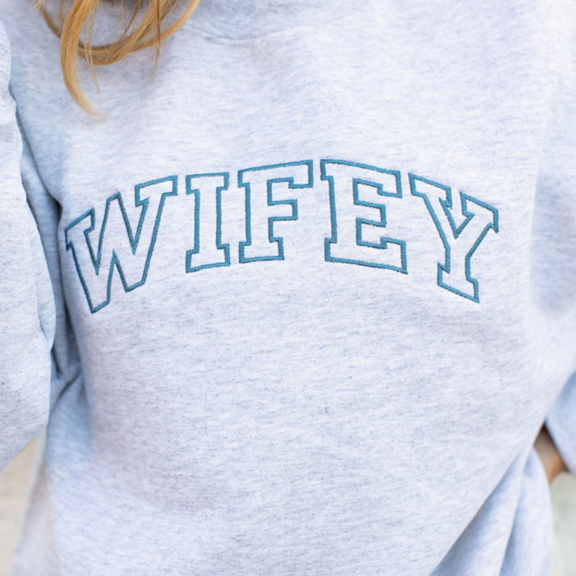close up of wifey embroidered in an athletic block font across the chest of a light gray crewneck pullover sweatshirt