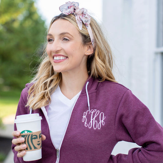 Blonde woman holding a travel coffee cup and wearing a heather maroon full zip jacket with script monogram in white thread 