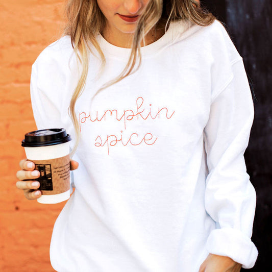 woman holding a coffee cup and wearing a white crewneck sweatshirt with the words pumpkin spice embroidered on the center of the sweatshirt in a chain stitch script font