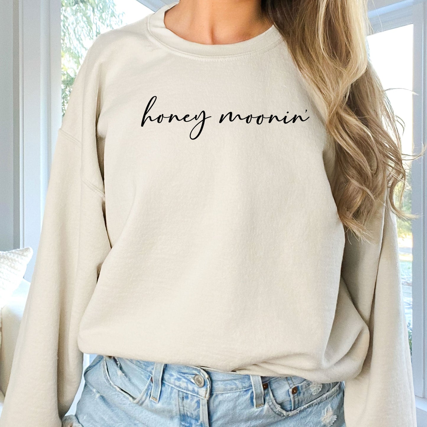 woman wearing a sand crewneck sweatshirt with honey moonin' printed across the chest in a black script font 
