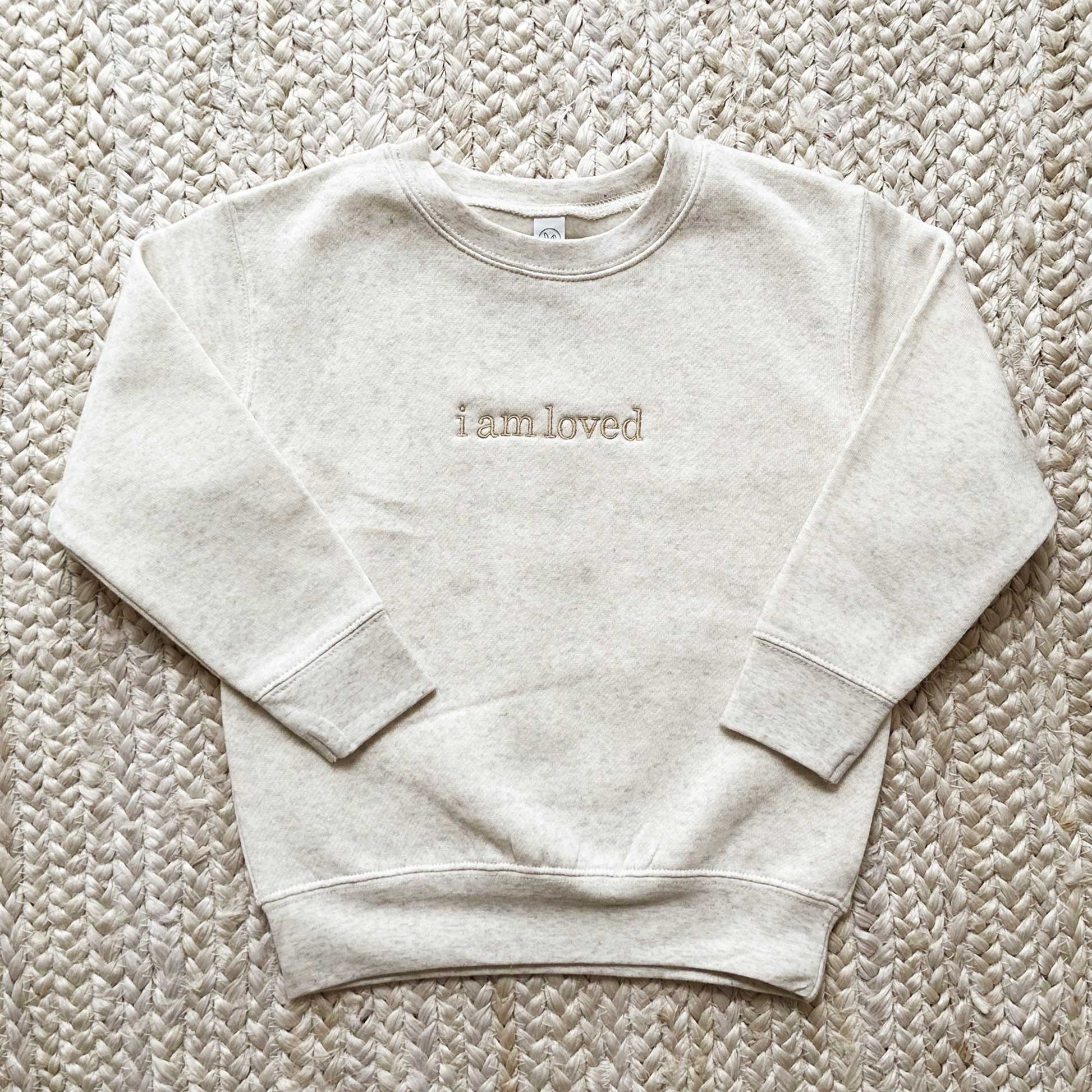 Toddler heather oatmeal crewneck sweatshirt with the words i am loved embroidered across the chest  in camel thread