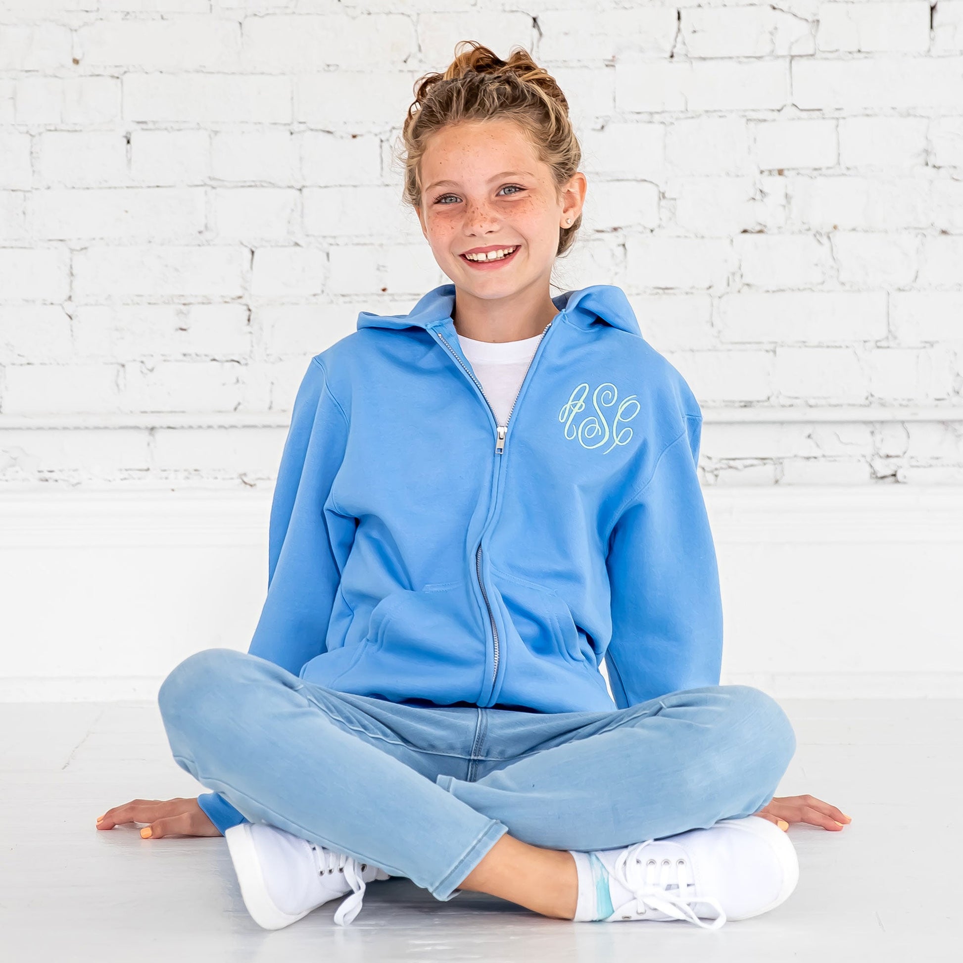 young girl sitting on the floor wearing jeans, sneakers, and a full zip hooded sweatshirt jacket with personalized monogram embroidery