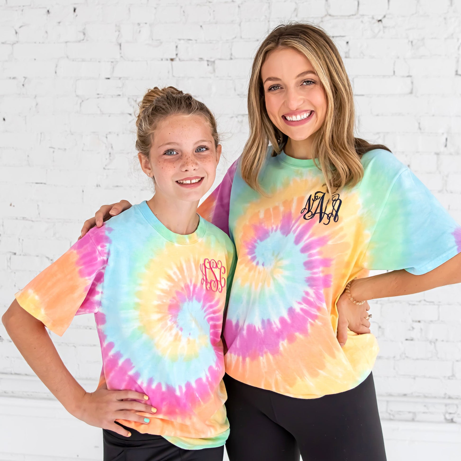girl and her big sister wearing matching t-shirts in a pastel rainbow tie dye with custom embroidered monograms on the left chest