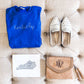 flatlay of outfit including a crossbody purse, white slides, and a royal blue crewneck with custom state stitched embroidery in white thread across the chest. 