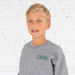 young boy wearing a heather gray pullover sweatshirt with custom monogram embroidery on the left chest in a forest green thread