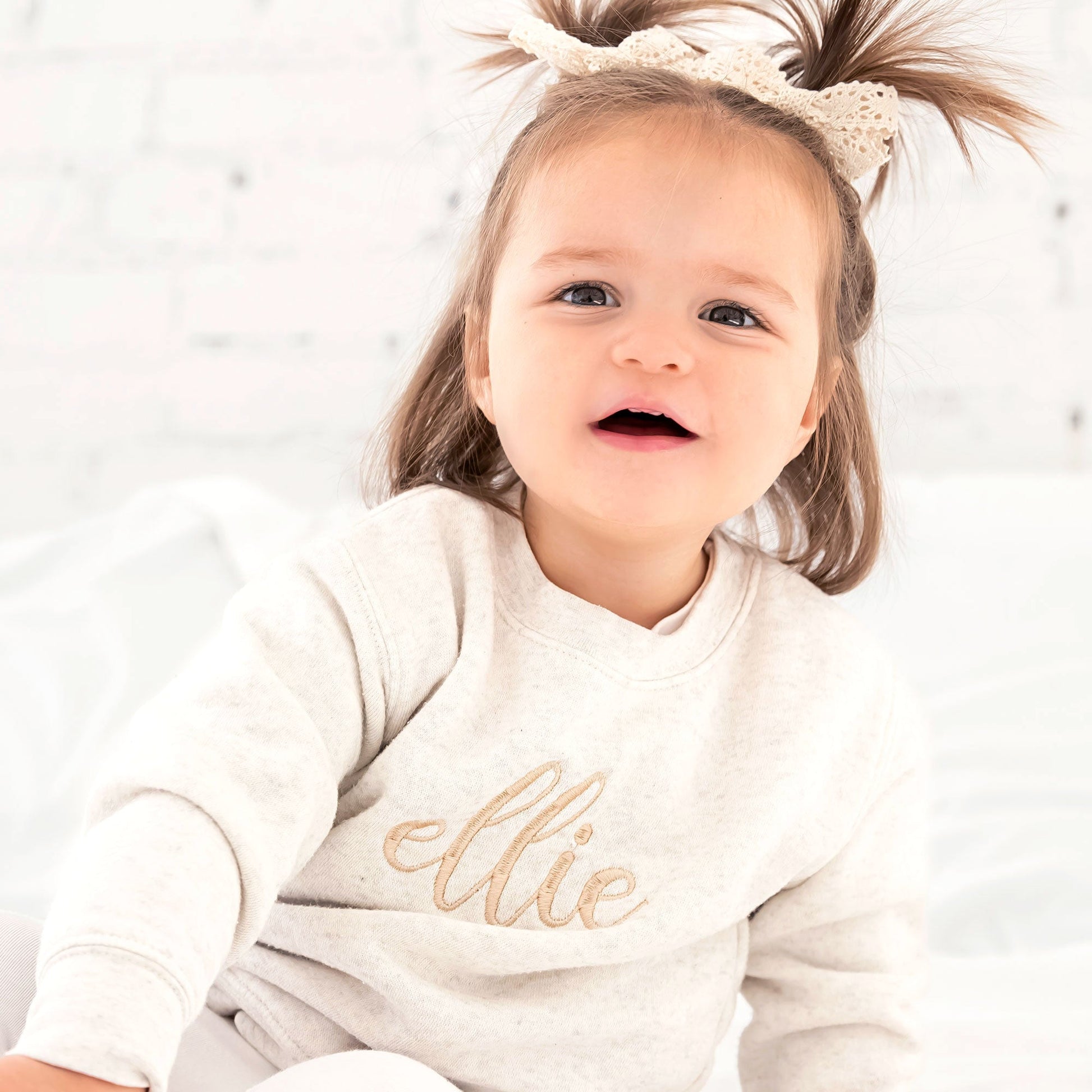 smiling little girl wearing an oatmeal crewneck sweatshirt with her name embroidered in a large script font across the front