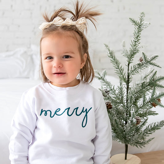 little girl sitting on a bench next to a miniature christmas tree and wearing a white crewneck sweatshirt with merry embroidered in a blue spruce green thread across the front
