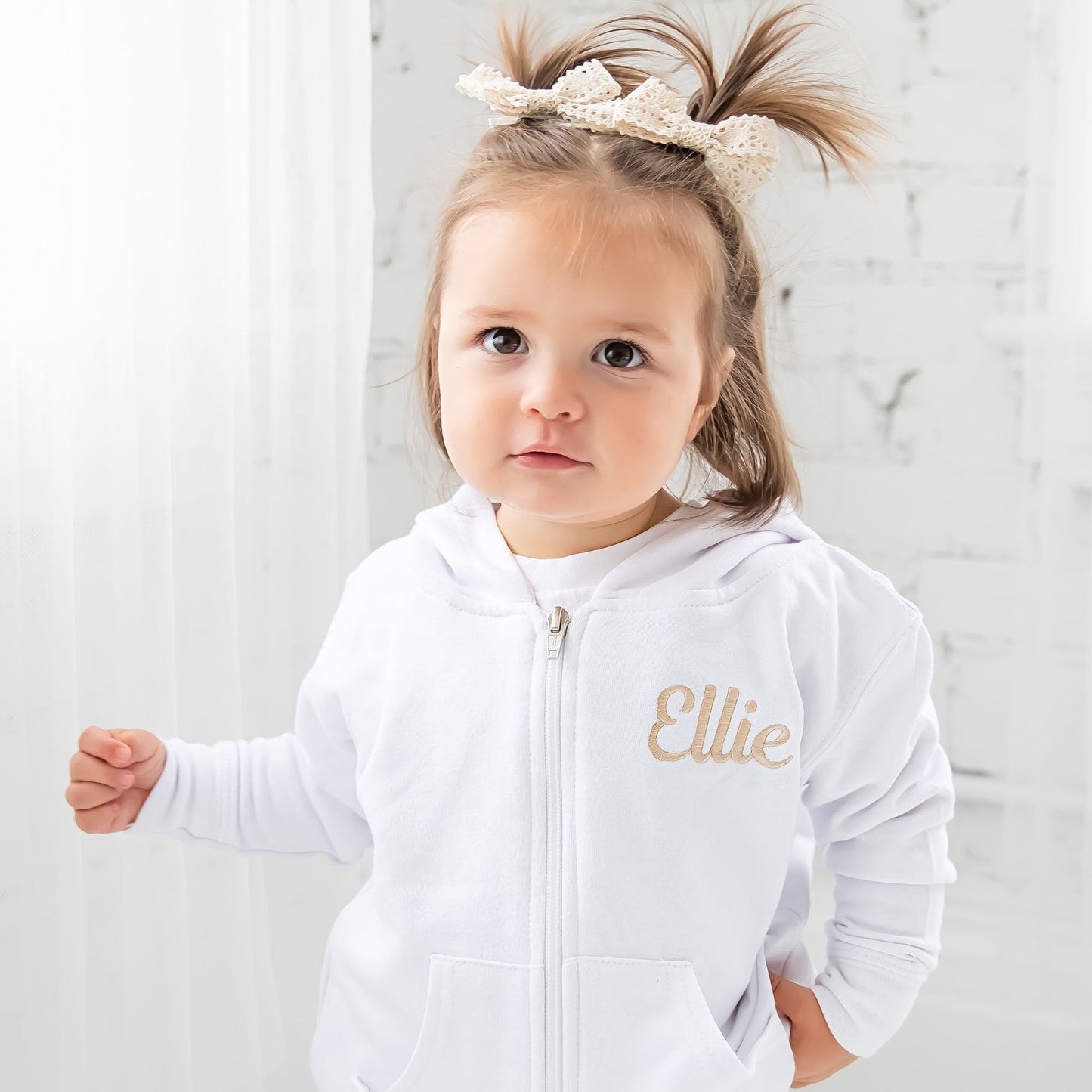 Little girl with pigtails wearing a white full zip hooded jacket featuring a custom name embroidery on the left chest in camel thread