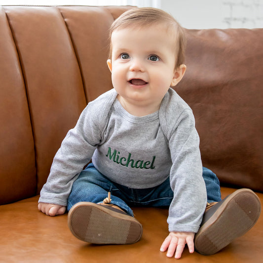 baby boy sitting on a couch wearing a pair of boots, jeans, and a long sleeve bodysuit with a custom name embroidery across the chest