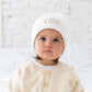 toddler wearing a cream colored cardigan and white beanie cap with her name embroidered in lowercase letters across the front cuff in camel thread