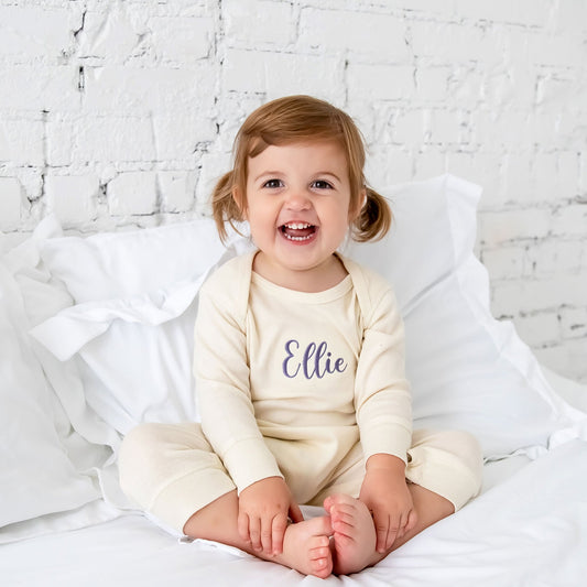 baby girl wearing a cream colored long sleeve bodysuit with a personalized name embroidery across the chest.