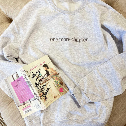 flatlay of a light gray crewneck sweatshirt with one more chapter embroidered across the chest in a dark gray thread
