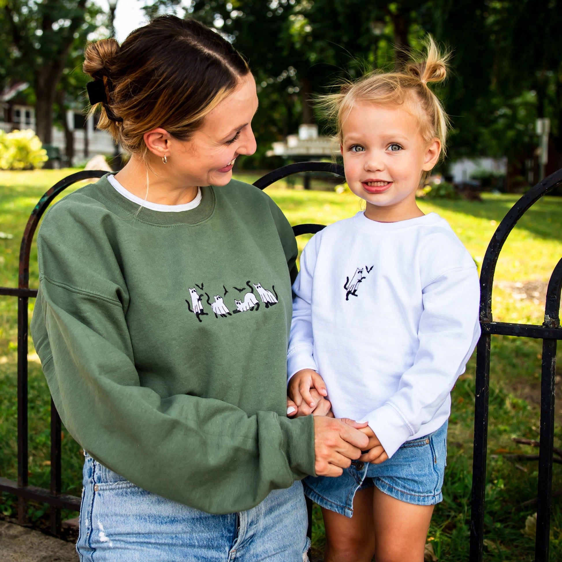 young woman wearing a military green crewneck sweatshirt with embroidered ghost cats design across the chest with her young daughter in a white crewneck sweatshirt with embroidered ghost cat