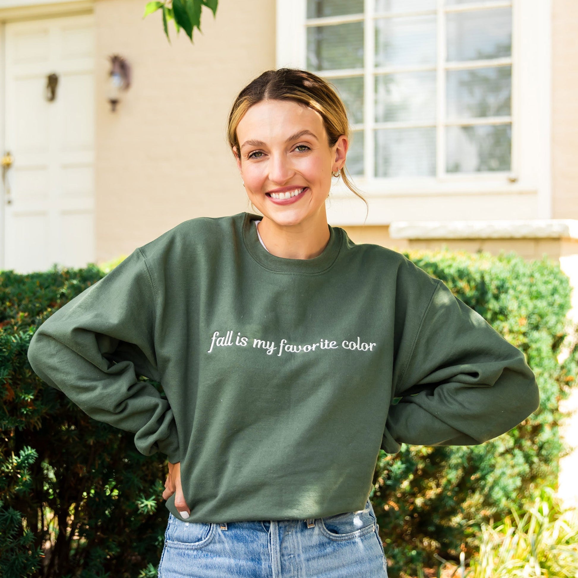 woman wearing jeans and a military green crewneck sweatshirt with a custom embroidery reading 'fall is my favorite color' in a natural thread color, standing outside a large cream brick house.