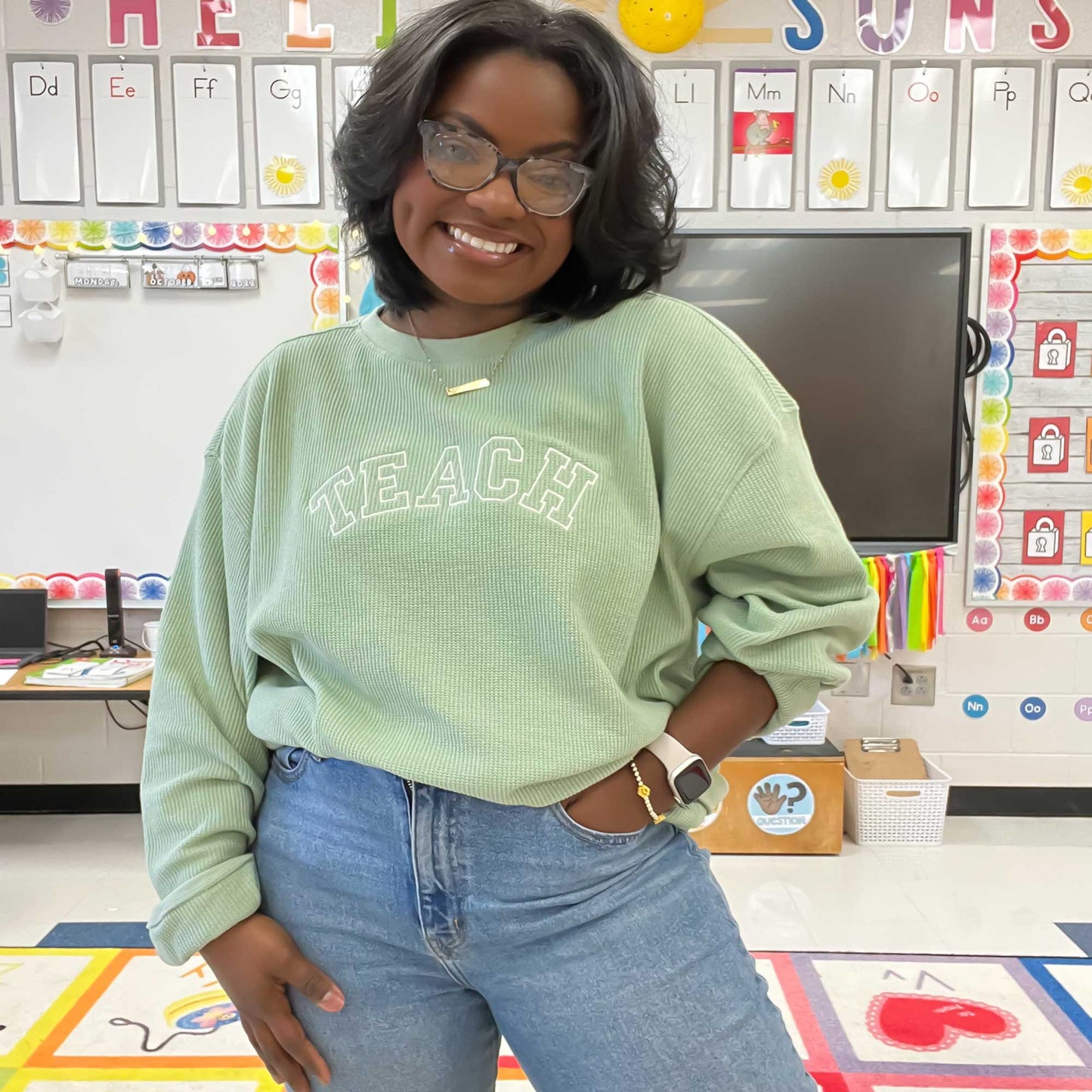 teacher in her classroom wearing a sage green waffle knit pullover with TEACH embroidered across the chest