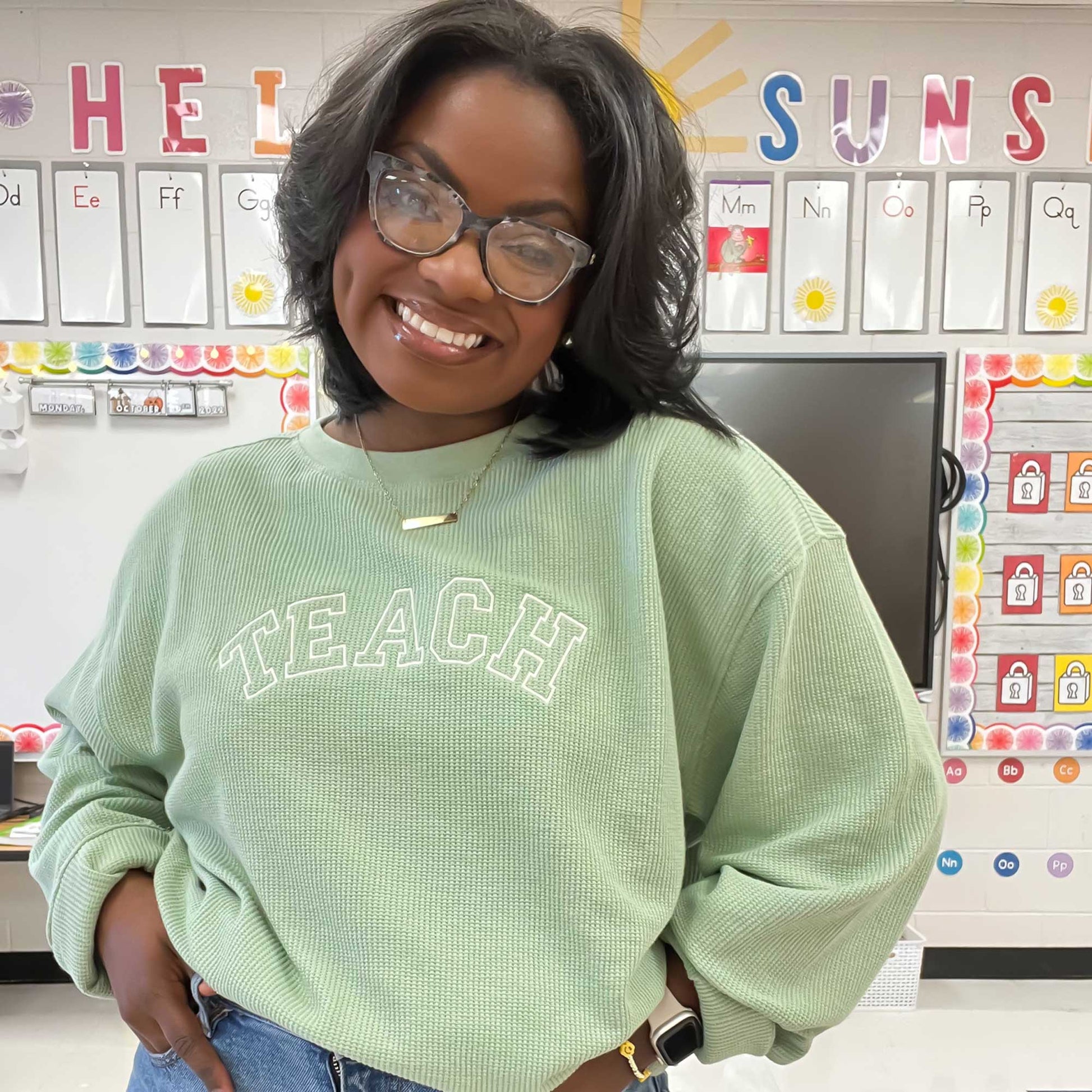 woman in her classroom wearing a sage waffle knit crew with embroidered TEACH design across the chest