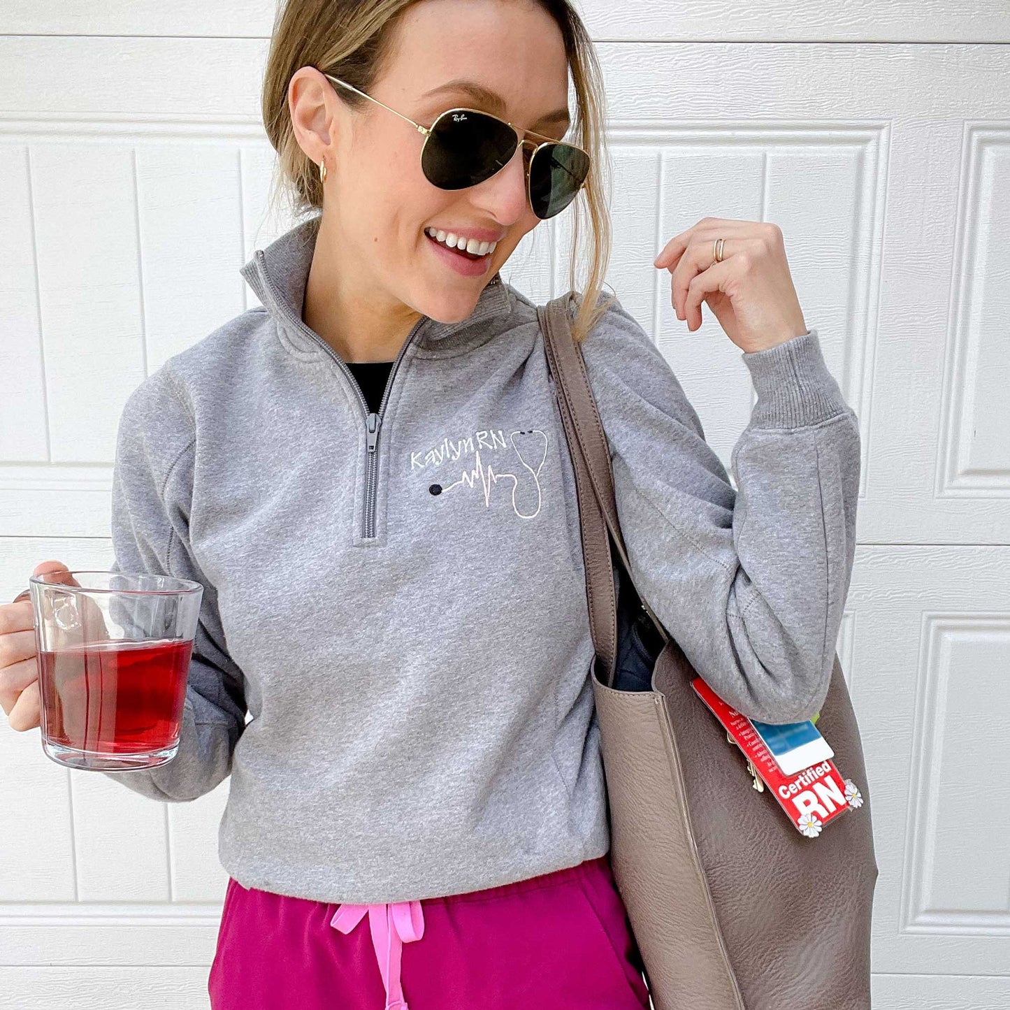 Nurse wearing pink scrub pants and a ladies fit vintage heather gray collared quarter zip with a custom name, credentials, and heartbeat stethoscope design embroidered on the left chest